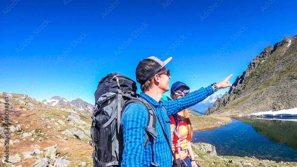 A couple with a big hiking backpacks, standing next to a clear, navy blue lake hiding between tall mountain peaks. Some of the slopes are covered with snow. In the back is another mountain range.