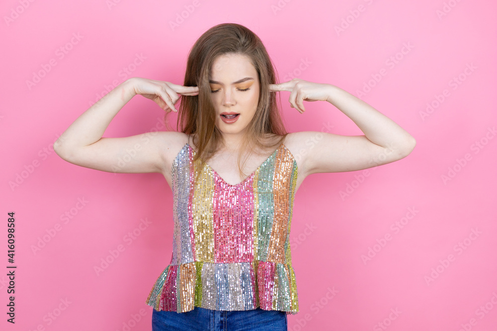 Pretty redhead woman wearing a shiny top standing over pink background covering ears with fingers with annoyed expression for the noise of loud music. Deaf concept. Pretty redhead woman wearing a shiny top standing over pink background covering ears with fingers with annoyed expression for the noise of loud music. Deaf concept.