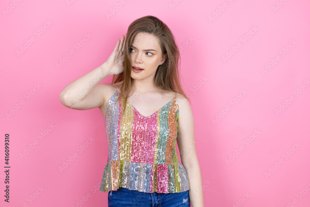 Pretty redhead woman wearing a shiny top standing over pink background surprised with hand over ear listening an hearing to rumor or gossip Pretty redhead woman wearing a shiny top standing over pink background surprised with hand over ear listening an hearing to rumor or gossip