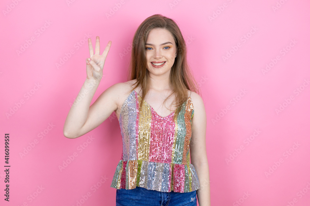 Pretty redhead woman wearing a shiny top standing over pink background showing and pointing up with fingers number three while smiling confident and happy Pretty redhead woman wearing a shiny top standing over pink background showing and pointing up with fingers number three while smiling confident and happy
