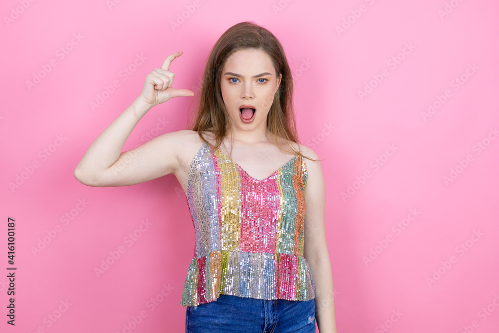 Pretty redhead woman wearing a shiny top standing over pink background smiling and confident gesturing with hand doing small size sign with fingers . Measure concept. Pretty redhead woman wearing a shiny top standing over pink background smiling and confident gesturing with hand doing small size sign with fingers . Measure concept.