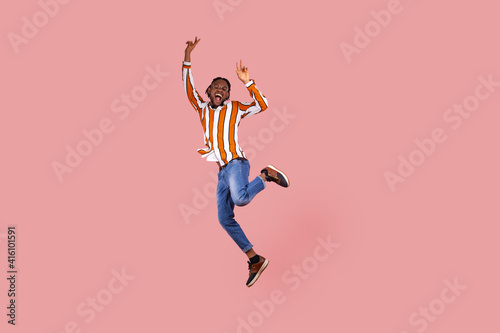 Full length excited ecstatic afro-american man with dreadlocks in striped shirt highly jumping posing, showing victory gesture, celebrating his triumph. Indoor studio shot isolated on pink background