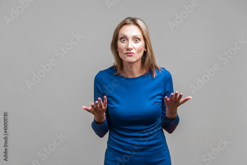 How could you? Portrait of disappointed unhappy woman in elegant tight blue dress standing with raised hands asking what do you want, angry face. indoor studio shot isolated on gray background