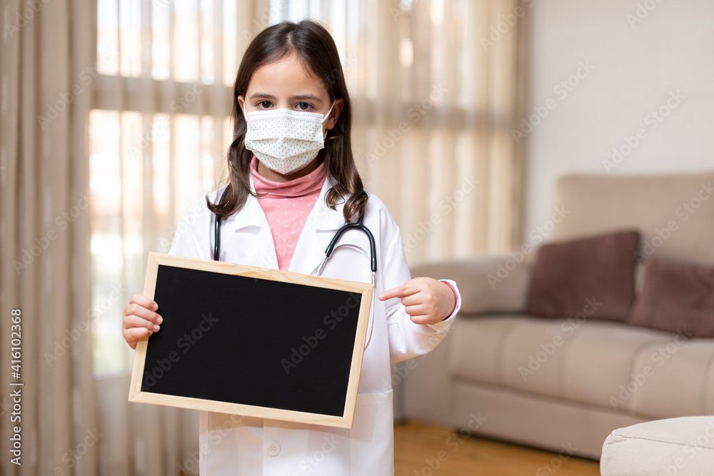 Little child dressed as a doctor and wearing a medical mask holding and pointing with her hand to a blackboard. She is at home. Space for text. Little child dressed as a doctor and wearing a medical mask holding and pointing with her hand to a blackboard. She is at home. Space for text.