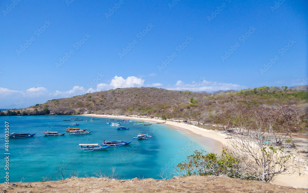 A view on a bay on Pink Beach, Lombok Indonesia. Plenty colourful boats anchored to the shore. The water has many shades of blue. The heavenly beach is surrounded by small hills. Paradise beach.