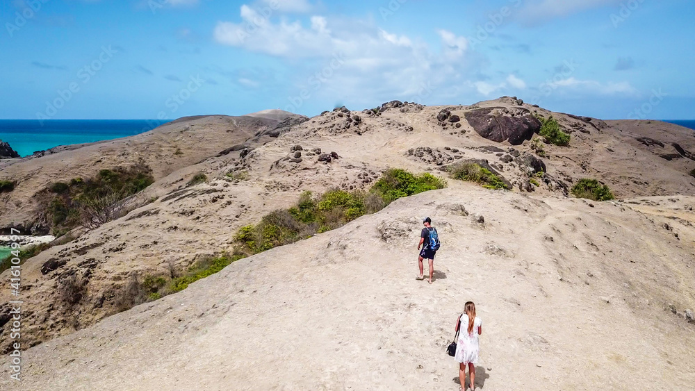 A drone shot of a couple walking on the cliffs, admiring the landscape in from of them. Overlooking famous Tanjung Aan beach on Lombok, Indonesia. Turquoise color of water. Happiness and togetherness