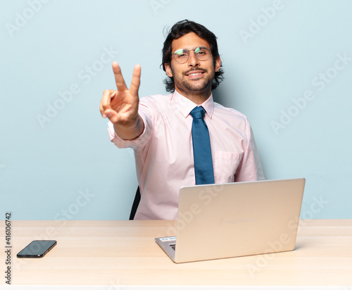 young handsome indian man smiling and looking happy, carefree and positive, gesturing victory or peace with one hand. business and frelancer concept photo