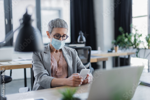 Mature businesswoman in medical mask using smartphone in office