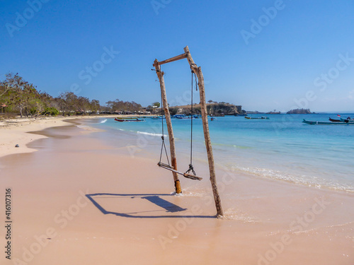 A swing placed on the seashore of Pink Beach  Lombok  Indonesia. The swing has very simple wood construction. Waves gently wash the pillars of it. In the back there are few boats anchored in the bay.