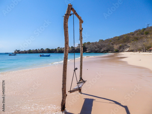 A swing placed on the seashore of Pink Beach, Lombok, Indonesia. The swing has very simple wood construction. Waves gently wash the pillars of it. In the back there are few boats anchored in the bay.