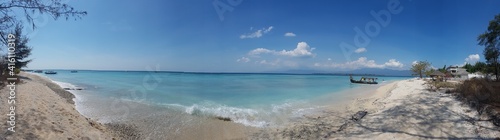 A panoramic shot of boats anchored next to the shore of Gili Air, Lombok Indonesia. Beautiful and clear water. In the back visible Mount Rinjani. Some trees on the shore, few clouds. Holidays paradise