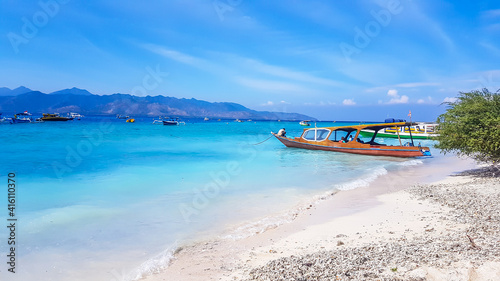Fototapeta Naklejka Na Ścianę i Meble -  A boat parked next to the shore on Gili Air, Lombok, Indonesia. Beautiful and clear water. In the back visible Mount Rinjani. Some trees on the shore, few clouds on the sky. Holidays paradise.