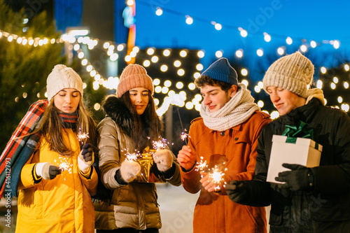 Group of four people student in warm clothes with burning sparklers or bengal lights enjoying parety in winter outdoors.