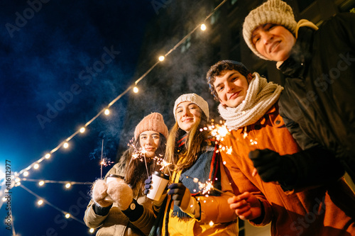Group of four people student in warm clothes with burning sparklers or bengal lights enjoying parety in winter outdoors.