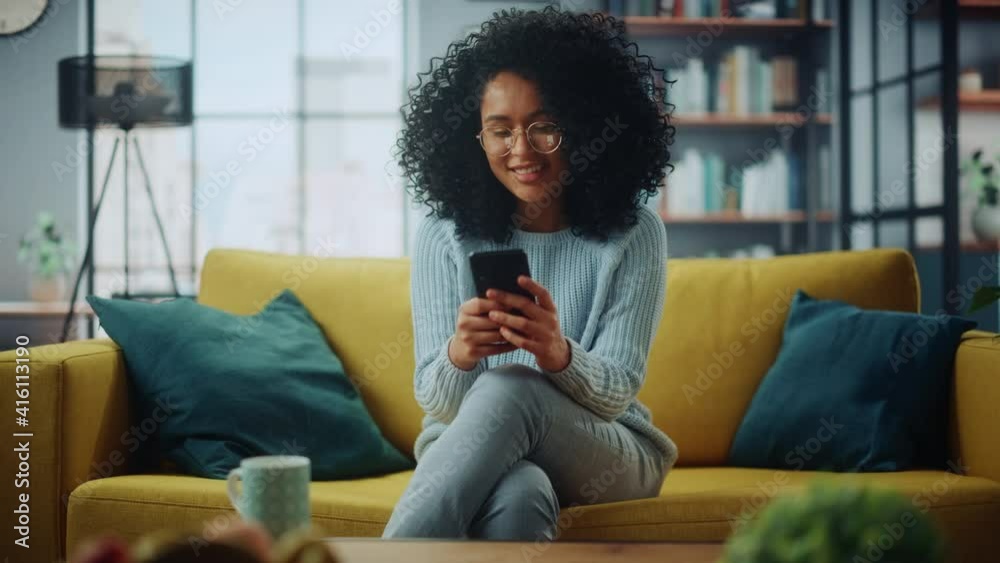 Portrait of a Beautiful Authentic Latina Female in a Stylish Cozy Living Room Using Smartphone at Home. She's Browsing the Internet and Checking Videos on Social Networks and Having Fun.