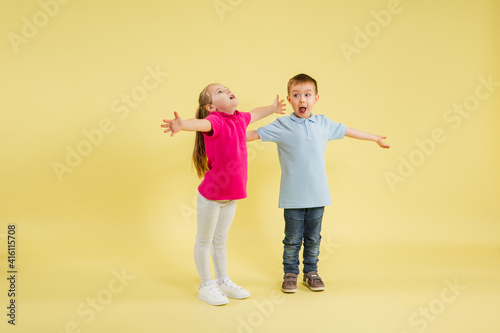 Greeting, showing. Childhood and dream about big and famous future. Pretty little kids isolated on yellow studio background. Dreams, imagination, education, facial expression, emotions concept.
