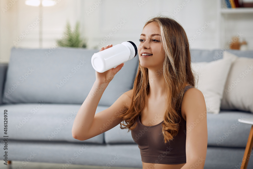 Athletic woman in sportswear sitting on the floor and drinking water from a bottle at home in the living room. Sport and recreation concept.
