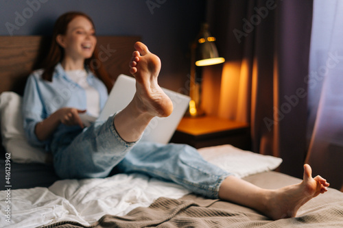 Cheerful redhead young woman chatting online via video call on laptop in bedroom lying at bed. Concept of leisure activity red-haired female at home during self-isolation.