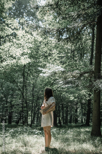 A young girl in a white dress stands in the forest with a basket in her hands and looks at the sun
