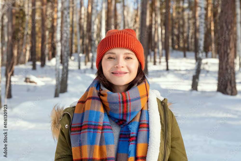 Young cheerful woman in warm jacket, checkered scarf and beanie looking at you