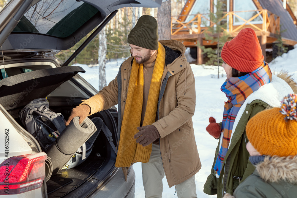 Young family of three in warm winterwear taking their buggage out of car trunk