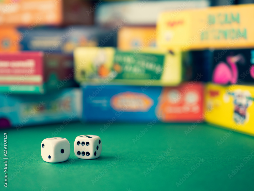 White dice on the green surface on the blurred background of colorful board game boxes. Concept of entertainment under lockdown - obrazy, fototapety, plakaty 