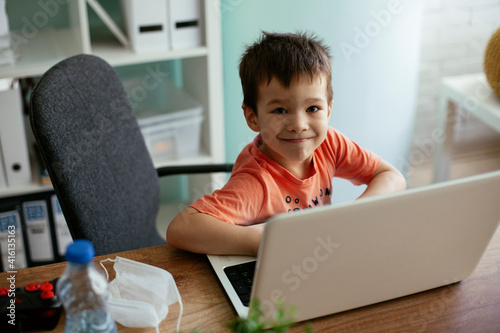 Portrait of cute smiling little boy sitting on office chair behind desk in the office. Happy boy using laptop.