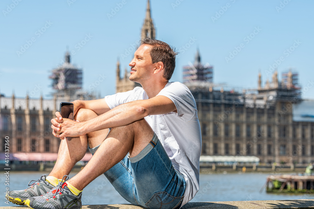 London, UK happy man sitting holding phone by cityscape Thames River Westminster during summer day on Albert Embankment and blue sky