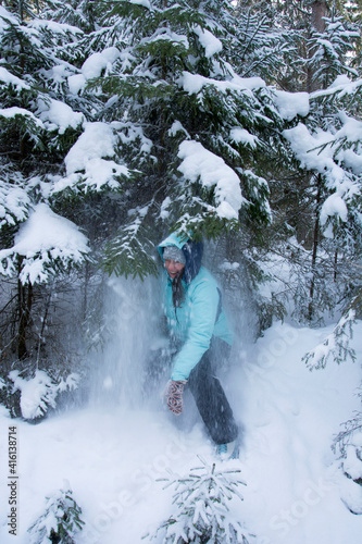 a girl in a turquoise sports jacket takes a snow shower from snow falling from a spruce tree