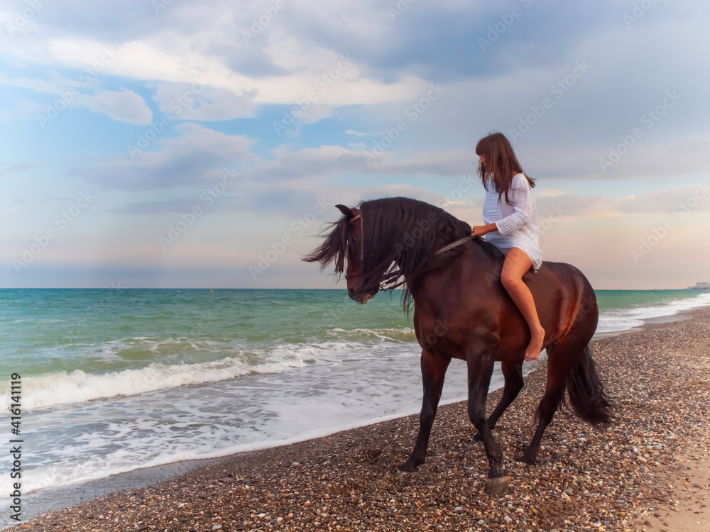 Joven amazona con vestido blanco domando a su caballo marron sin montura en la playa Joven amazona con vestido blanco domando a su caballo marron sin montura en la playa