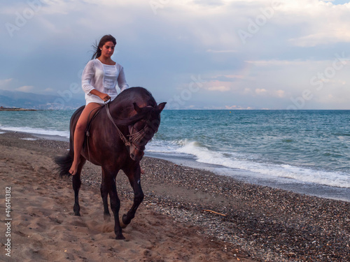 Joven amazona con vestido blanco domando a su caballo marron sin montura en la playa