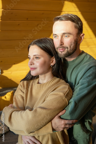 Young affectionate husband embracing wife while standing in front of window