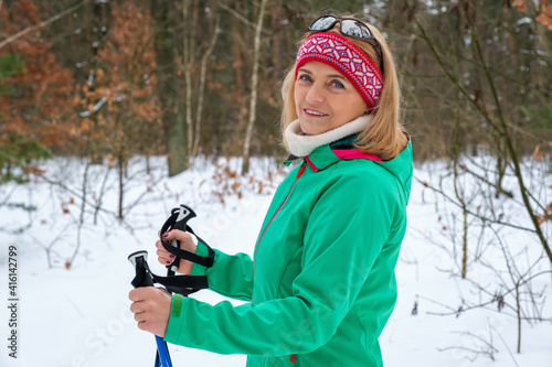 Elderly woman standing with nordic walking poles in winter forest, resting after exercise and looking at camera outdoors with smile.