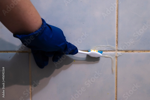 A hand in a blue glove cleans the dirty joints between the tiles in the bathroom with an old toothbrush. Toxic black mold in the bathroom during the cleaning process.