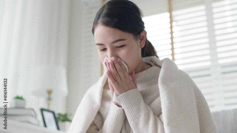 Sick young asian woman sitting under the blanket on sofa and sneeze with tissue paper at home. Female blowing nose, coughing or sneezing in tissue at home, suffering from flu. Cold and fever concept Sick young asian woman sitting under the blanket on sofa and sneeze with tissue paper at home. Female blowing nose, coughing or sneezing in tissue at home, suffering from flu. Cold and fever concept