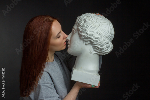 Red-haired young girl on black background in a gray T-shirt kissing with a plaster bust