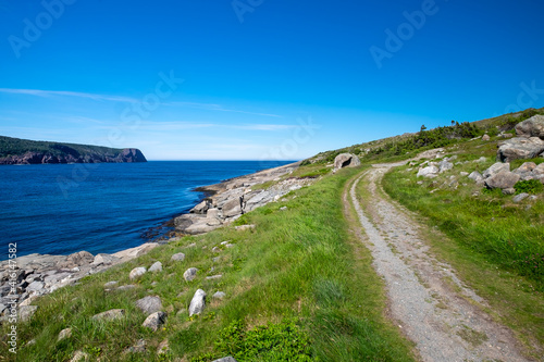 Worn gravel and rocky footpath or hiking trail along a coastline near the edge of blue ocean's water in a cove. There's a tree covered island in the distance near the horizon with a deep blue sky. 