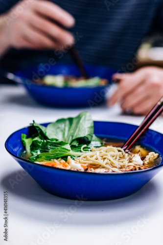 Ramen noodles with egg and bok choy in a cobalt blue bowl, man eating in the background