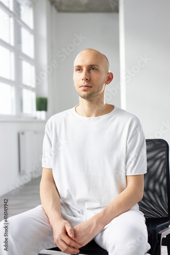 yogi male in white clothing sit in contemplation having rest, calm young man with bald head engaged in yoga, sport. isolated in bright studio room