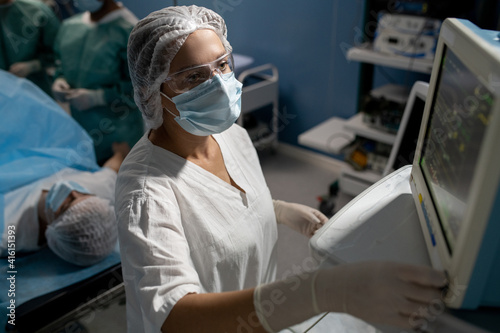 Serious assistant of surgeon in gloves, mask and uniform looking at display