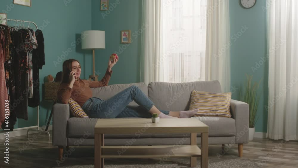 Beautiful girl in blue jeans and a brown sweater lies on a beige sofa on the background of the window, holding a red apple and talking on the phone. General plan.