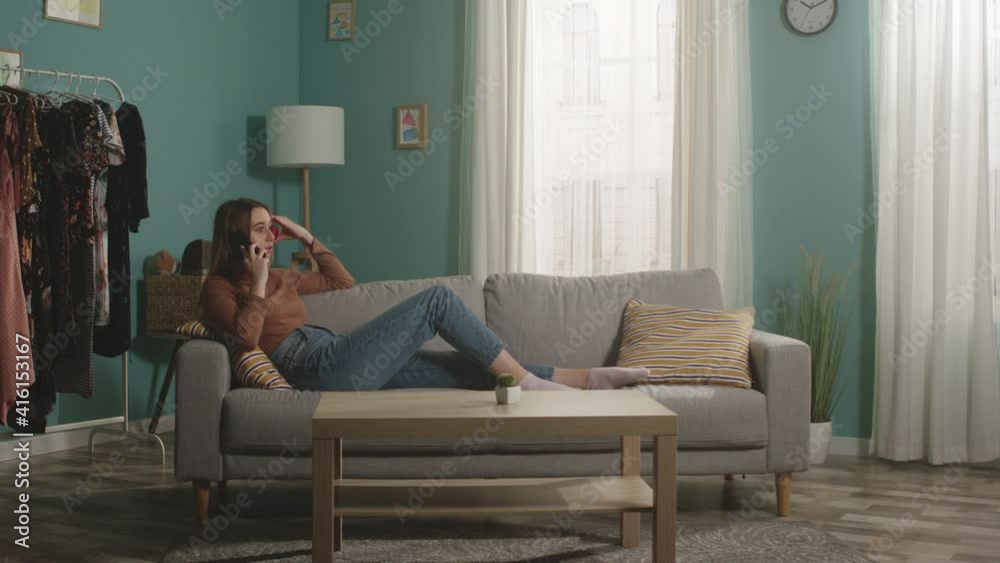 Young lady in blue jeans and a brown sweater lies on a beige sofa on the background of the window, holding a red apple and talking on the phone. General plan. Young lady in blue jeans and a brown sweater lies on a beige sofa on the background of the window, holding a red apple and talking on the phone. General plan.
