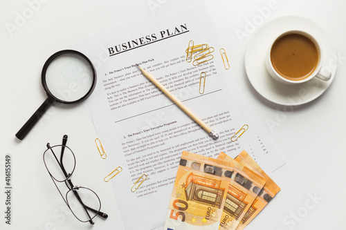 Flat lay of business plan documents with money, glasses, coffee cup, paper clip and supplies on white table desk