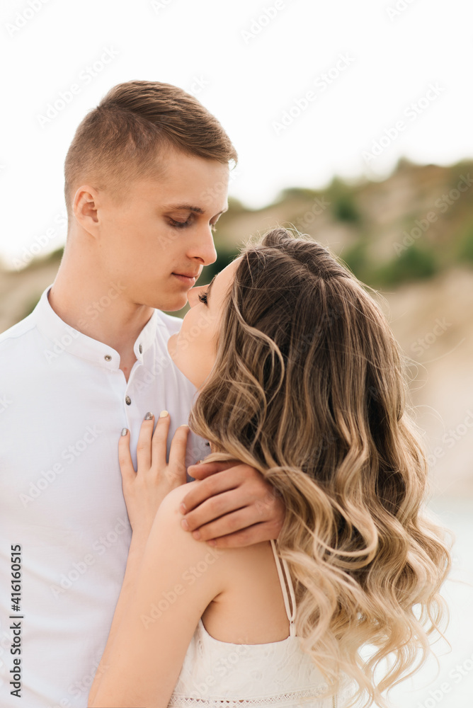 Fototapeta premium A beautiful young couple in love, a man and a woman embrace, kiss near a blue lake and sand at sunset. Vacation at the sea on the beach, sunset, morning, honeymoon, tropics, ocean, girl, relationship.