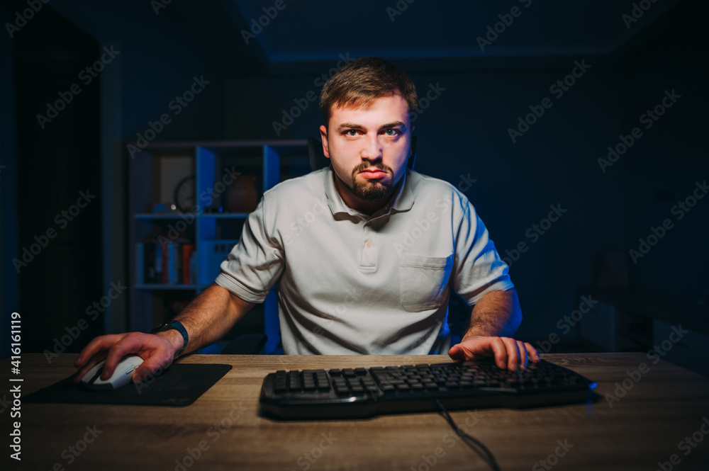 Serious bearded freelancer works at home on the computer at night and looks at the camera with a serious face. Freelance programmer works online at home. Serious bearded freelancer works at home on the computer at night and looks at the camera with a serious face. Freelance programmer works online at home.
