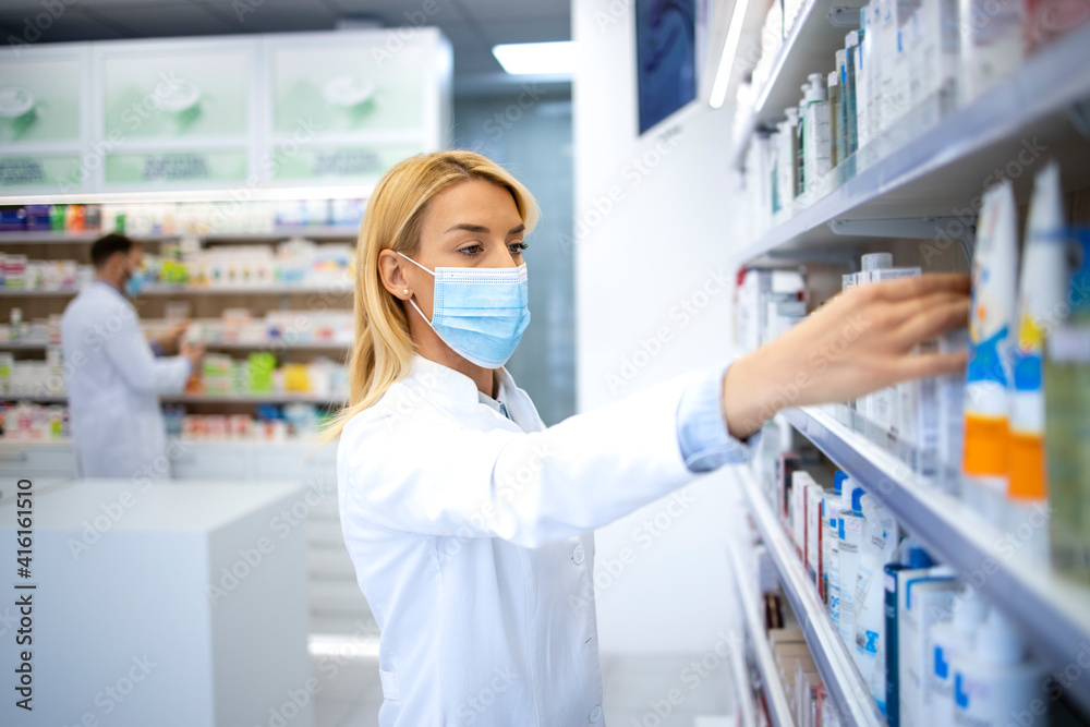 Female pharmacist wearing face mask and white coat working in pharmacy store during corona virus pandemic.
