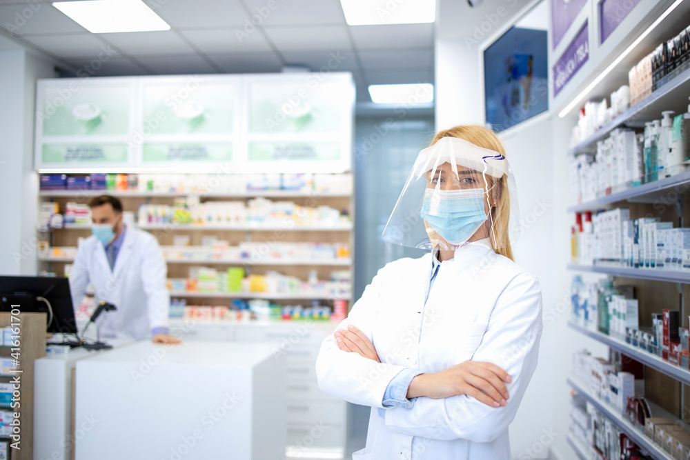 Portrait of female pharmacist wearing face shield and white coat standing in pharmacy store during corona virus pandemic.