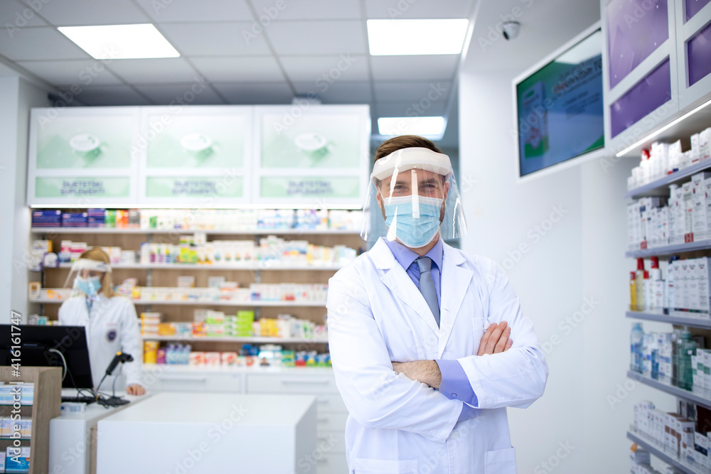 Portrait of pharmacist wearing face shield and white coat standing in pharmacy store during corona virus pandemic.