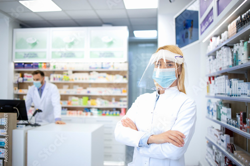 Portrait of female pharmacist wearing face shield and white coat standing in pharmacy store during corona virus pandemic.