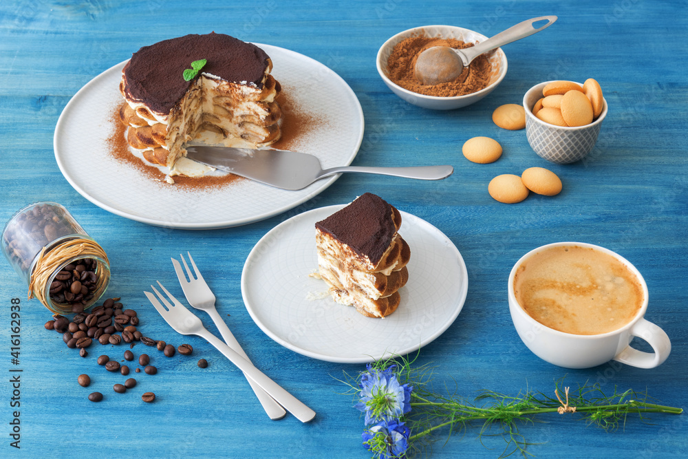 Round tiramisu, homemade no-bake traditional Italian sweet dessert. Blue table with cake, bowl with cocoa powder, round sponge biscuits, coffee beans and coffee with milk. Blue background. Round tiramisu, homemade no-bake traditional Italian sweet dessert. Blue table with cake, bowl with cocoa powder, round sponge biscuits, coffee beans and coffee with milk. Blue background.